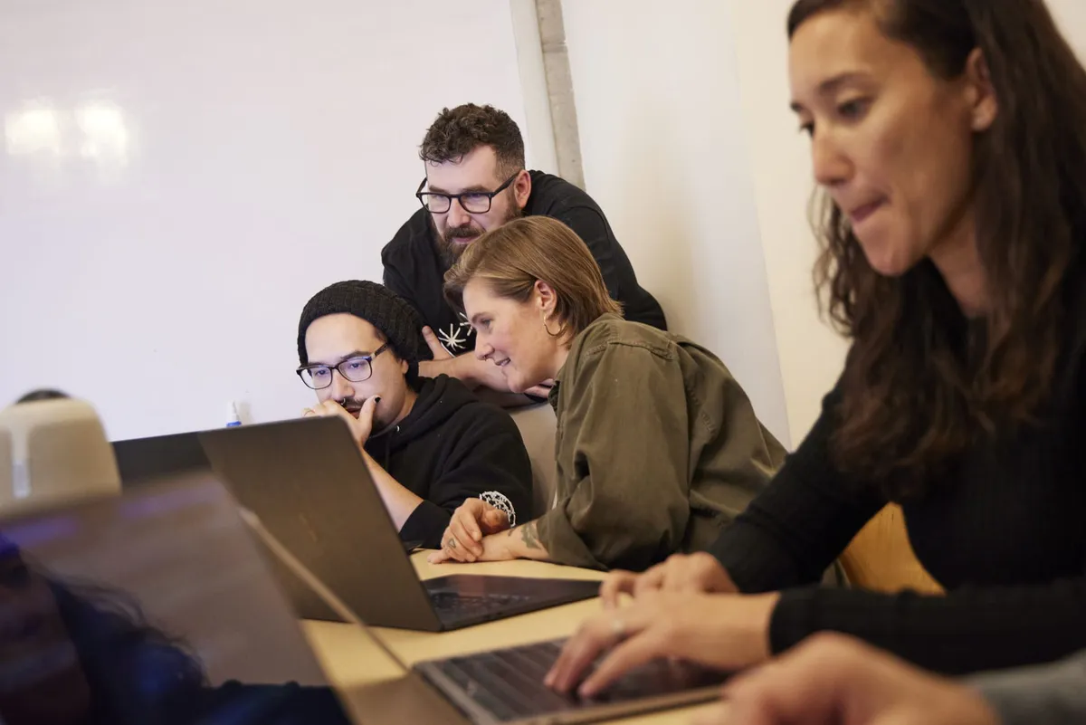A woman works on her laptop in the foreground while three colleagues gather around another screen behind her.