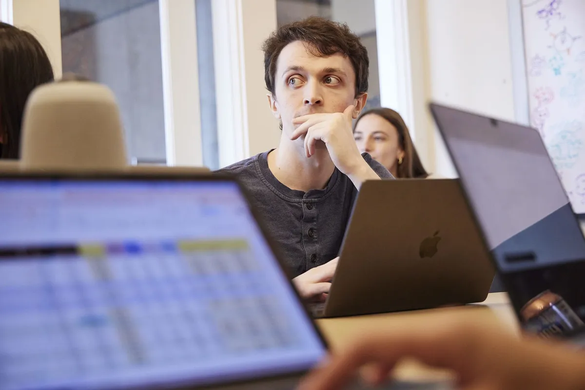 A man rests his chin on his hand, thinking at a laptop in a bright office, with a blurred spreadsheet in the foreground.