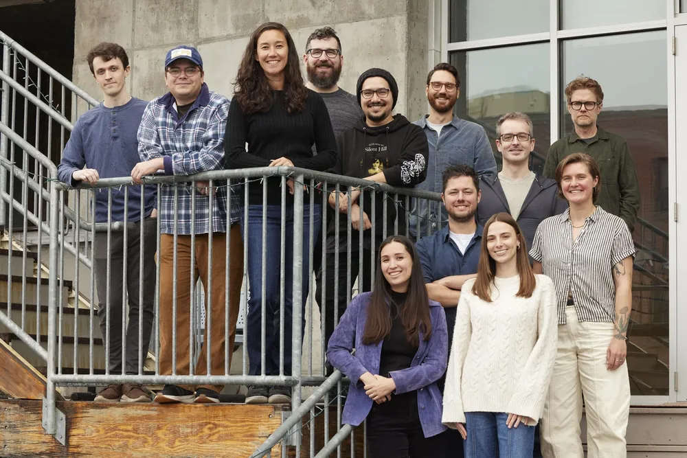 Looking up at 12 members of the Bluehouse Group team, smiling and standing on a staircase. A few are leaning on the handrail.