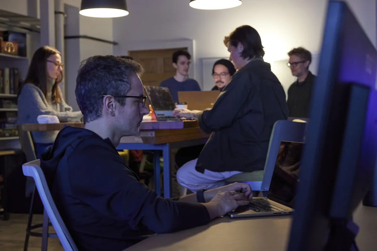 A man focuses on his laptop while colleagues collaborate around a table in the background