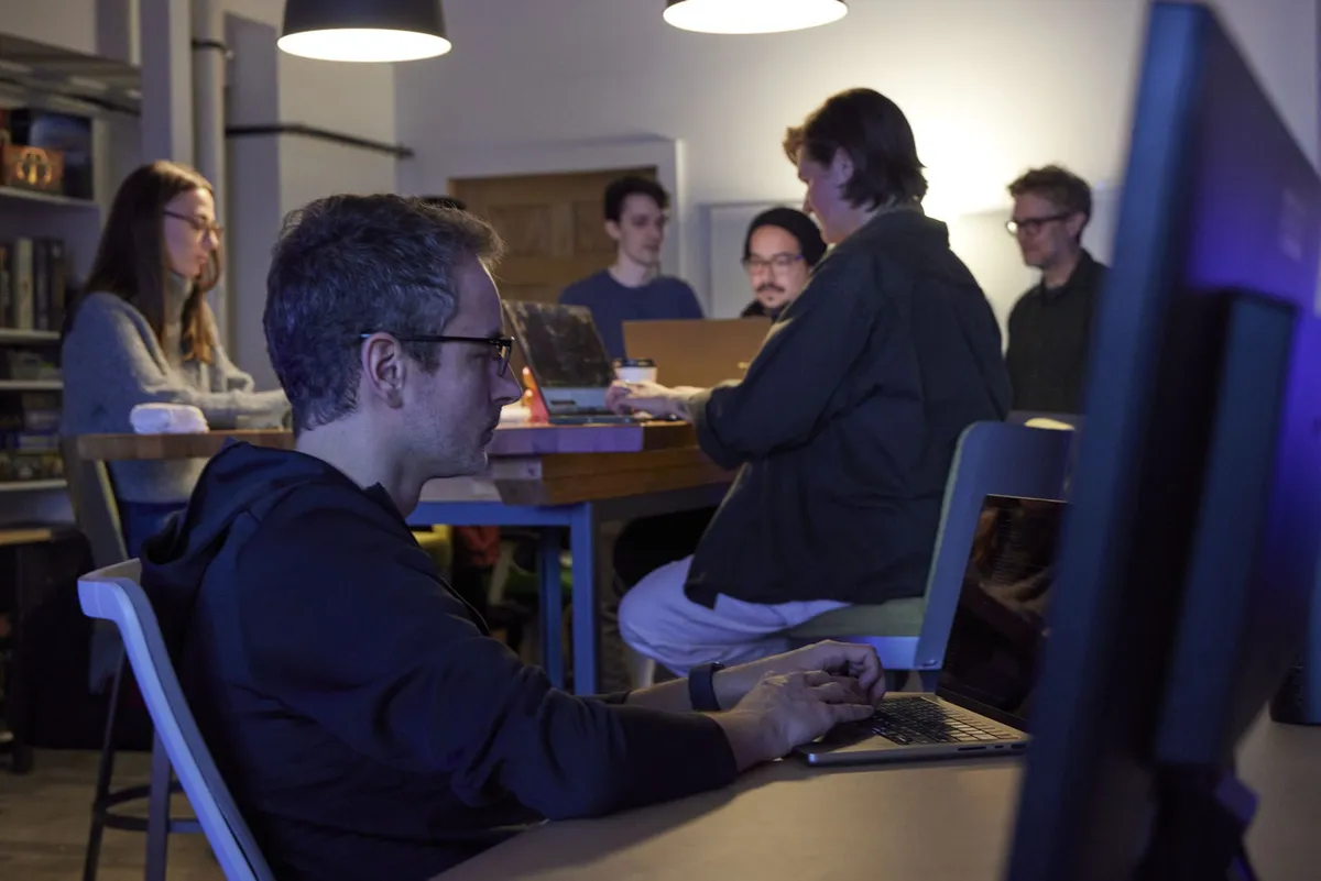 A man focuses on his laptop while colleagues collaborate around a table in the background