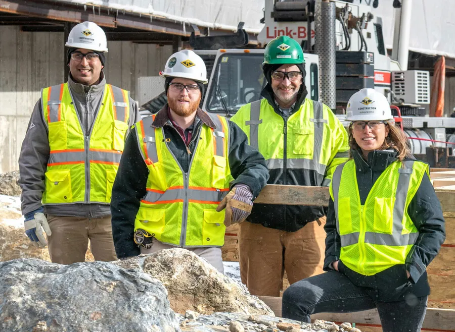 Four construction workers in safety vests and hard hats smile at a job site next to construction equipment and boulders.