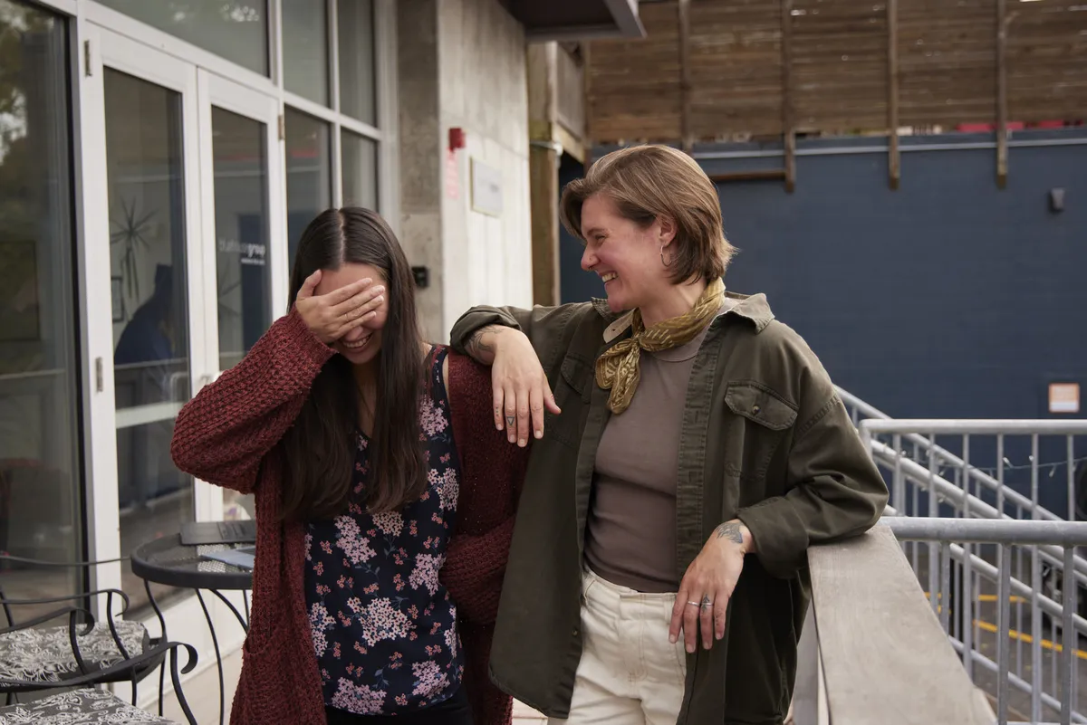 Two friends sharing a laugh, one leaning on the other’s shoulder while the other covers their face