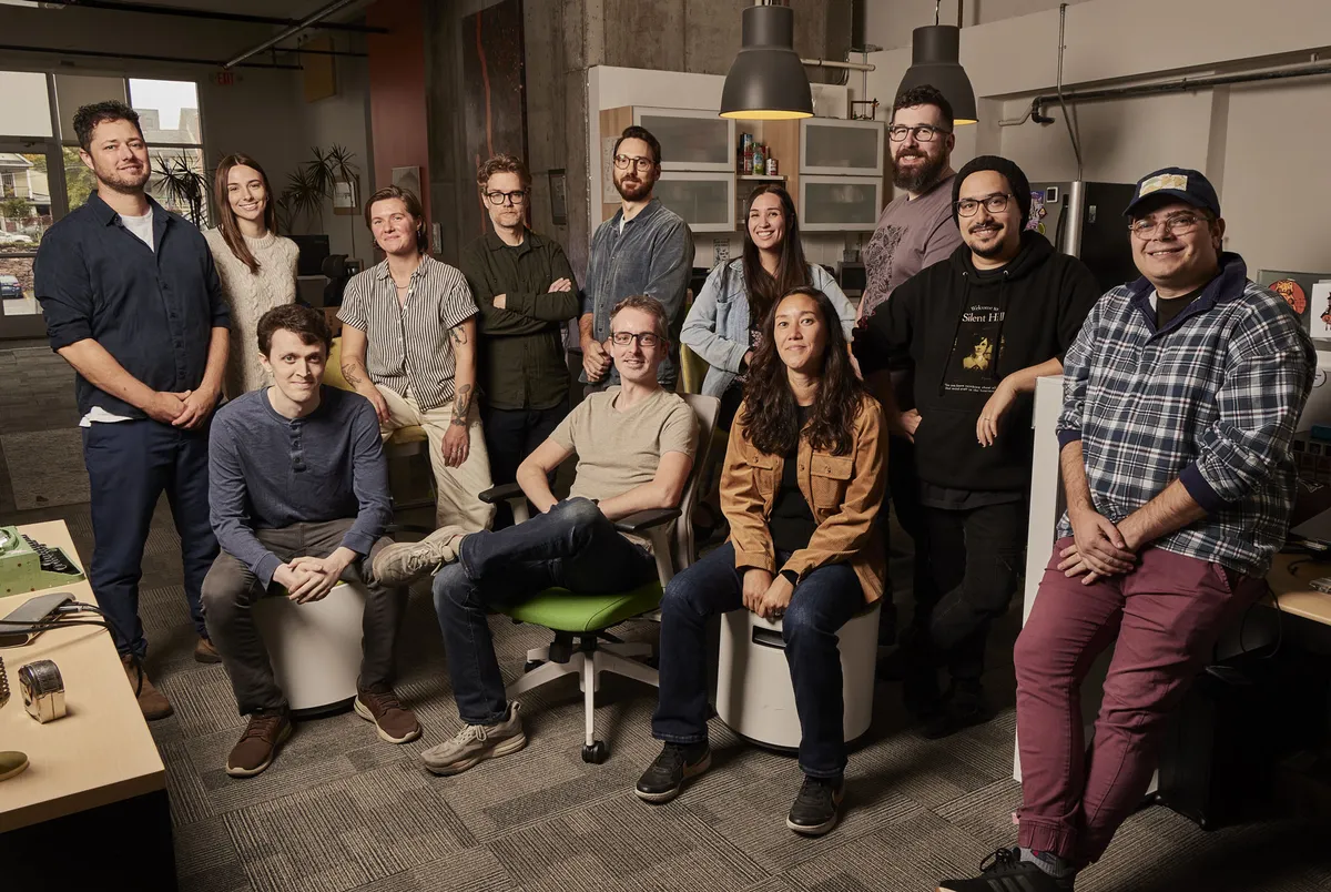 A group photo of 12-member team in the office, with some members seated on stools or chairs and others standing. Everyone is smiling, except for one person who refuses to smile. The office features exposed concrete walls, large windows, and various office equipment.