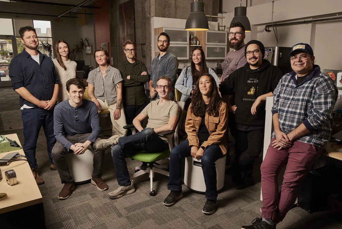 A group photo of 12-member team in the office, with some members seated on stools or chairs and others standing. Everyone is smiling, except for one person who refuses to smile. The office features exposed concrete walls, large windows, and various office equipment.