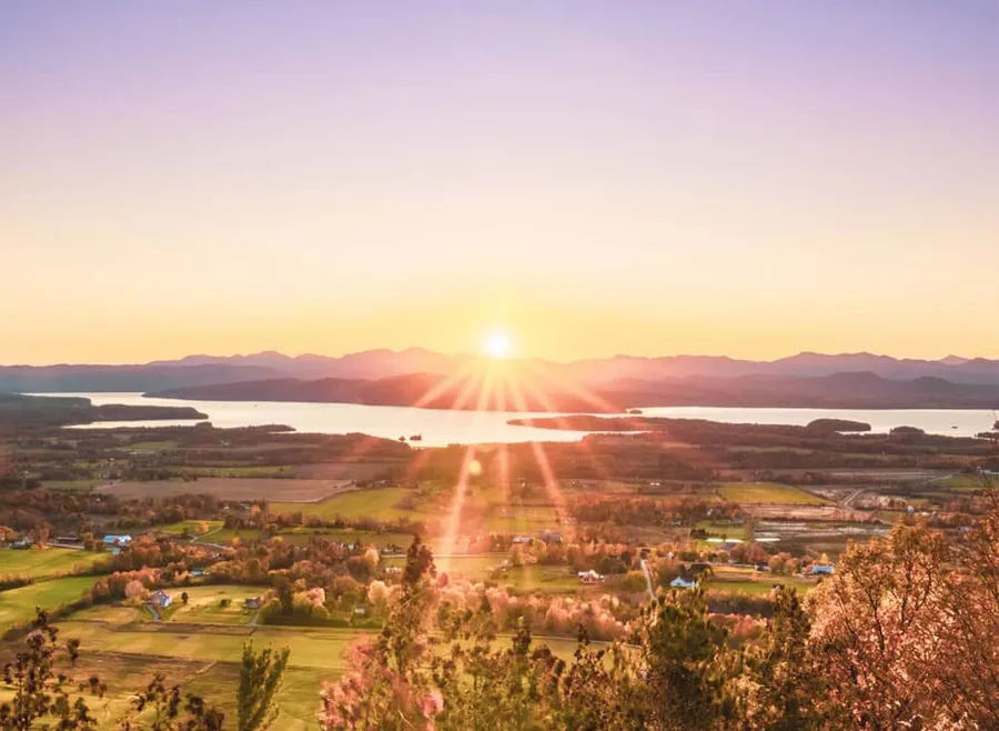 View from Mt. Philo, the sun is setting over the Adirondack Mountains and Lake Champlain, with farmland in the valley below.
