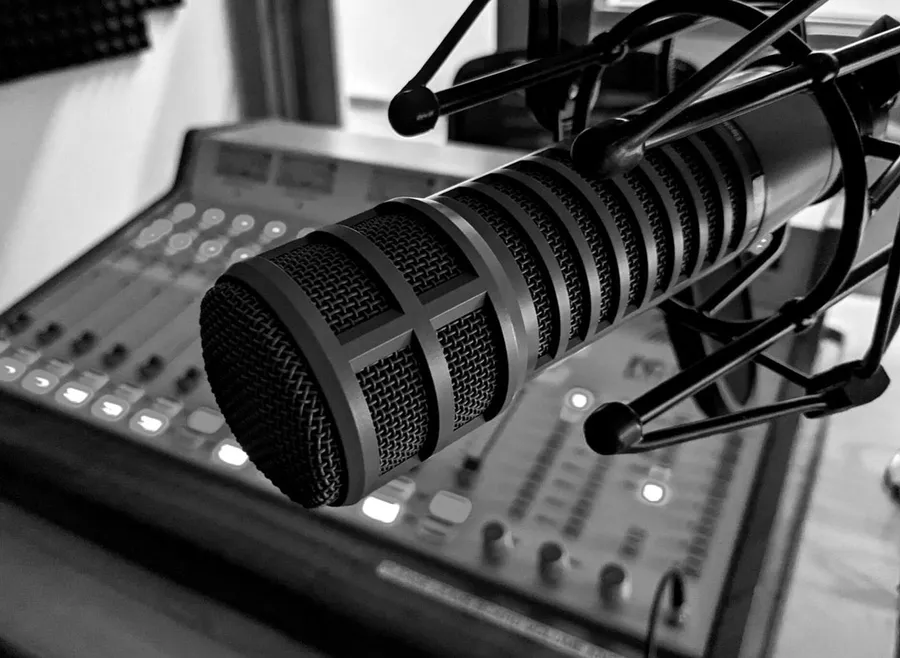 Microphone on a stand positioned above a sound mixing board in a radio studio. The mixing board in the background is slightly out of focus.