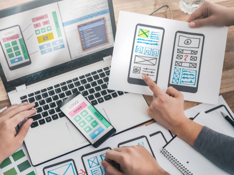 A group of people working on a mobile app design, you can only see their hands. They are using a laptop, and multiple sheets of paper with low fidelity drawings of mobile app screens on the papers. 