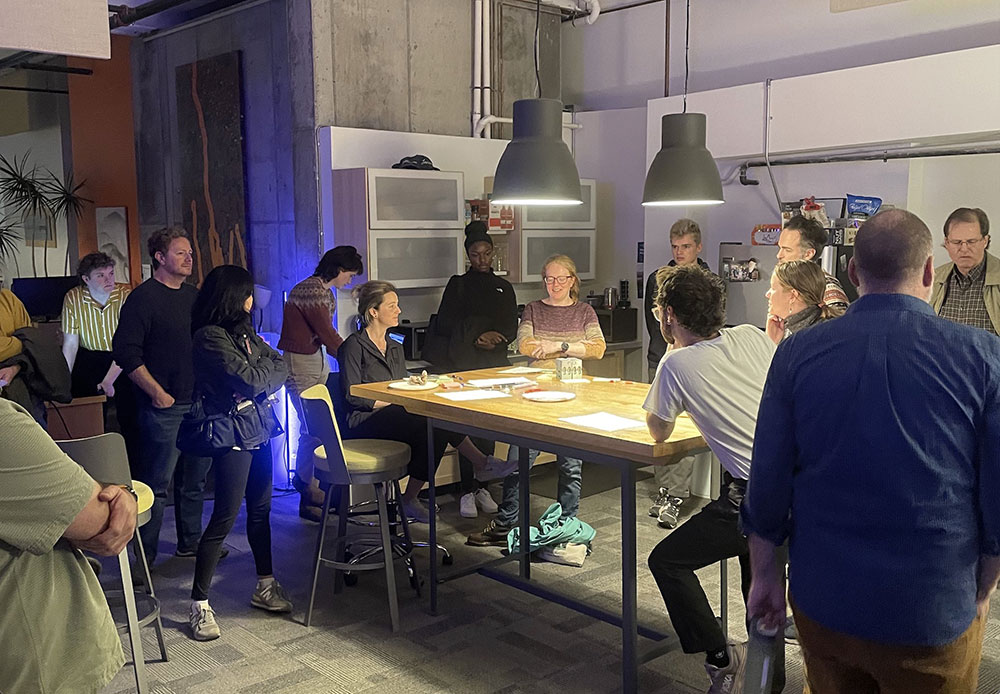 A large group of people are standing and sitting around a table. Everyone in the group is listening to a women presenting at the center of the table. There are papers, markers, pens, and sticky notes on the table. While not visible in the image, the papers contain notes, ideas, and low fidelity wireframes with UX and UI solutions for an Open Data Portal for the city of Burlington, the women is presenting these ideas. 